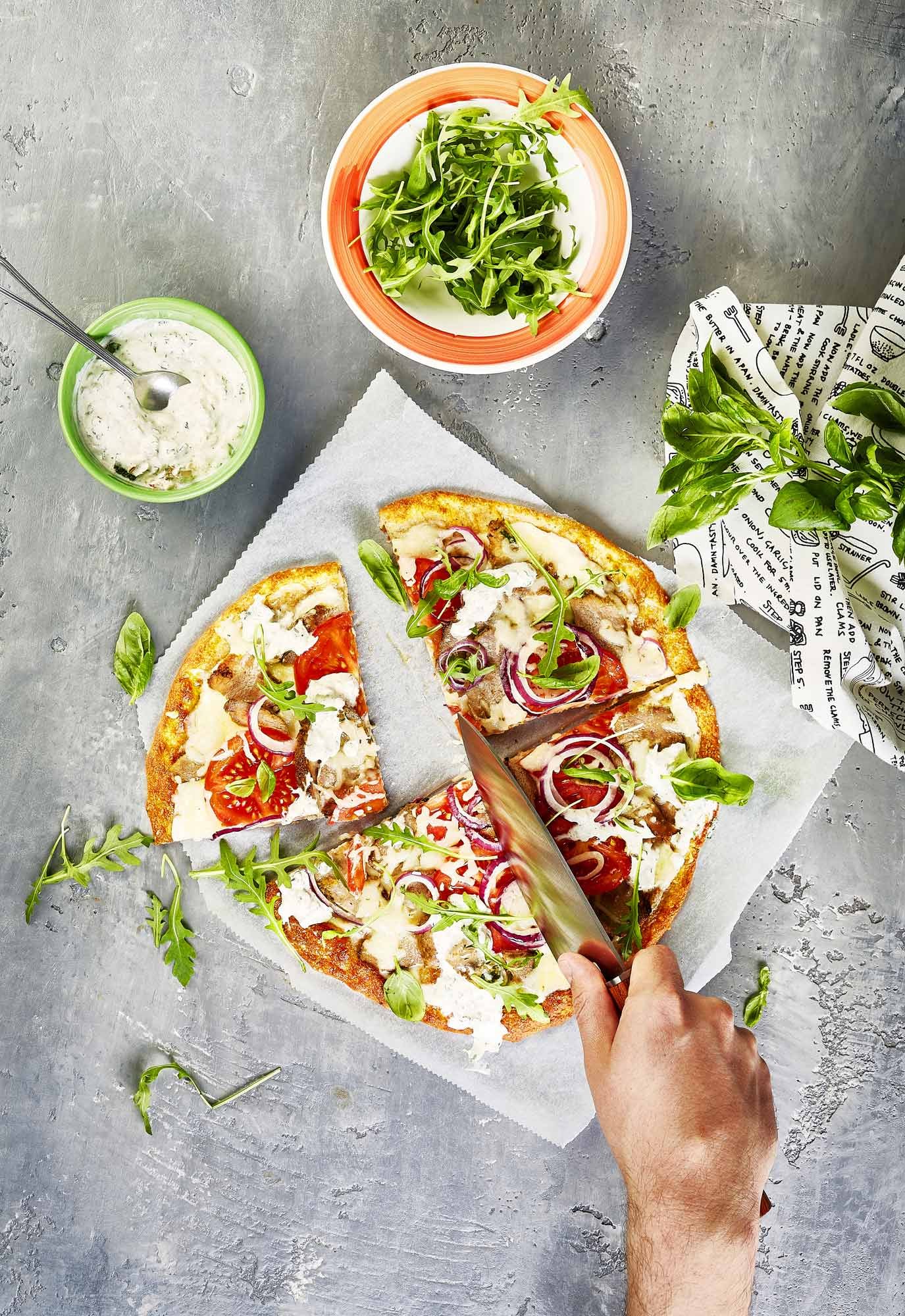 photographie culinaire en top shot, main coupant une pizza cétogène au kebab et à la roquette